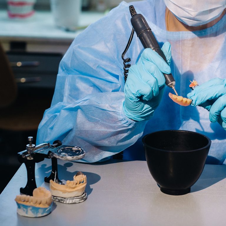 A masked and gloved dental technician works on a prosthetic tooth in his lab.